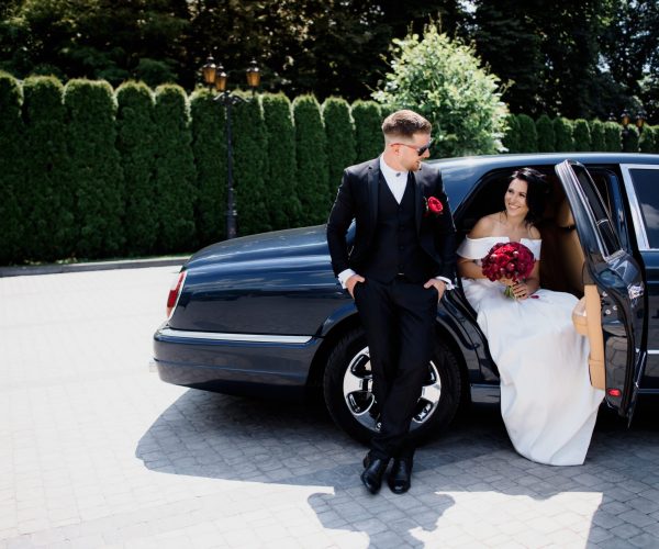 Beautiful wedding couple is smiling in the black car on the sunn Beautiful wedding couple is smiling in the black car on the sunny day, dressed in elegant wedding outfits with red bouquet